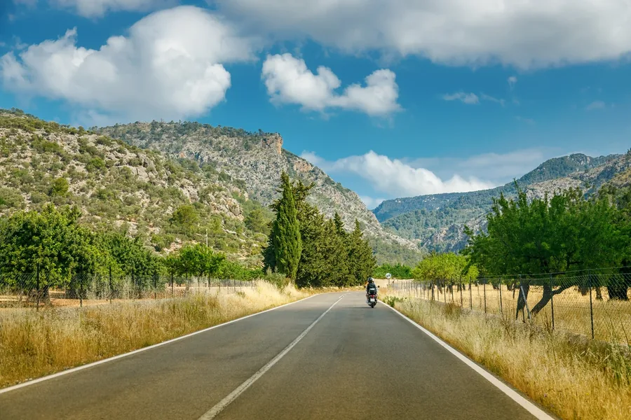 Motorrad mit Topcase auf einer leeren Landstraße auf Mallorca, dahinter Zypressen und Berge