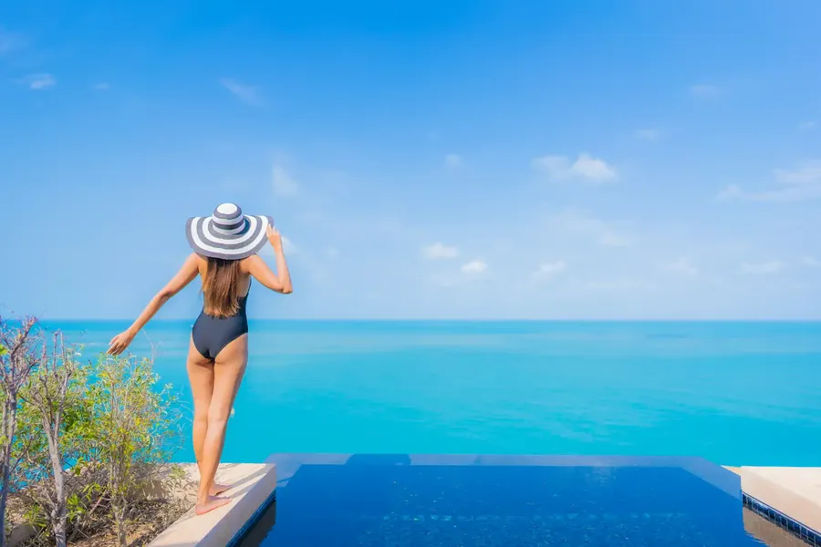 Eine Frau mit gestreiftem Sonnenhut blickt über einen Pool aufs Meer